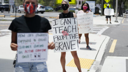 Teachers hold signs standing on a sidewalk. One in focus reads 