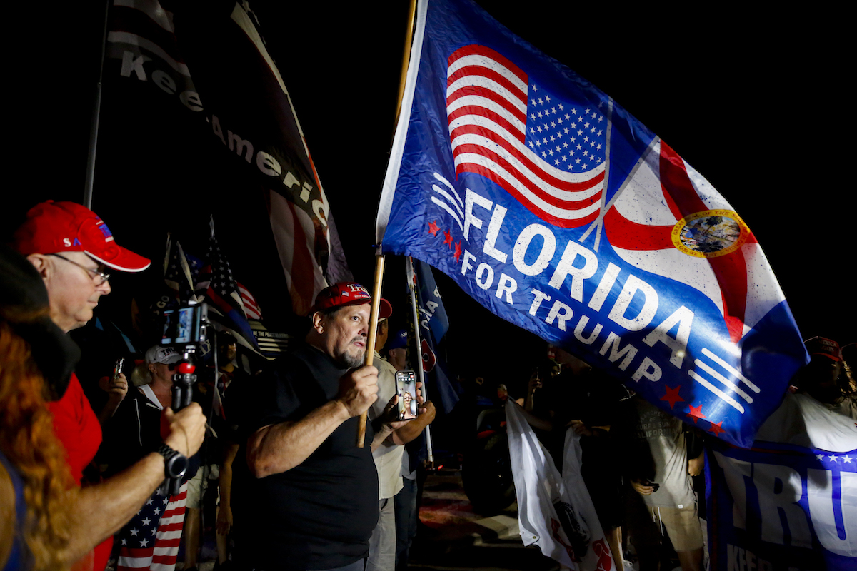 Trump's supporters protest outside at night, holding flags reading "Florida for Trump"