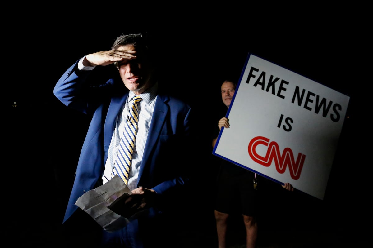 A man holding a sign that reads Fake News is CNN stands behind a news anchor waiting to go on-air.