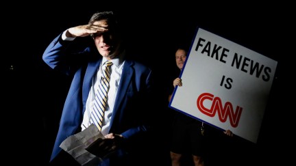 A man holding a sign that reads Fake News is CNN stands behind a news anchor waiting to go on-air.