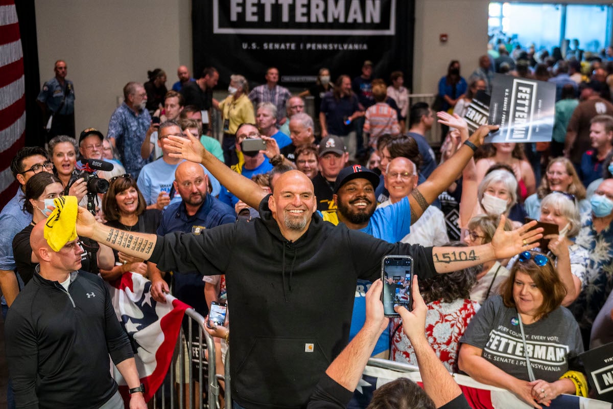John Fetterman spreads his arms wide and grins standing in front of a crowd of supporters