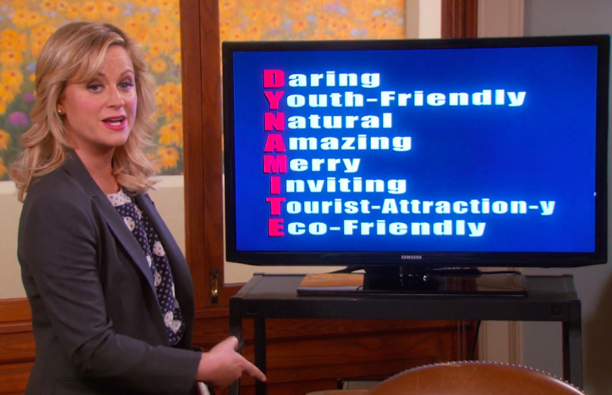 Leslie Knope stands in front of a presentation screen with acrostic "DYNAMITE" displayed in a scene from Parks & Recreation.