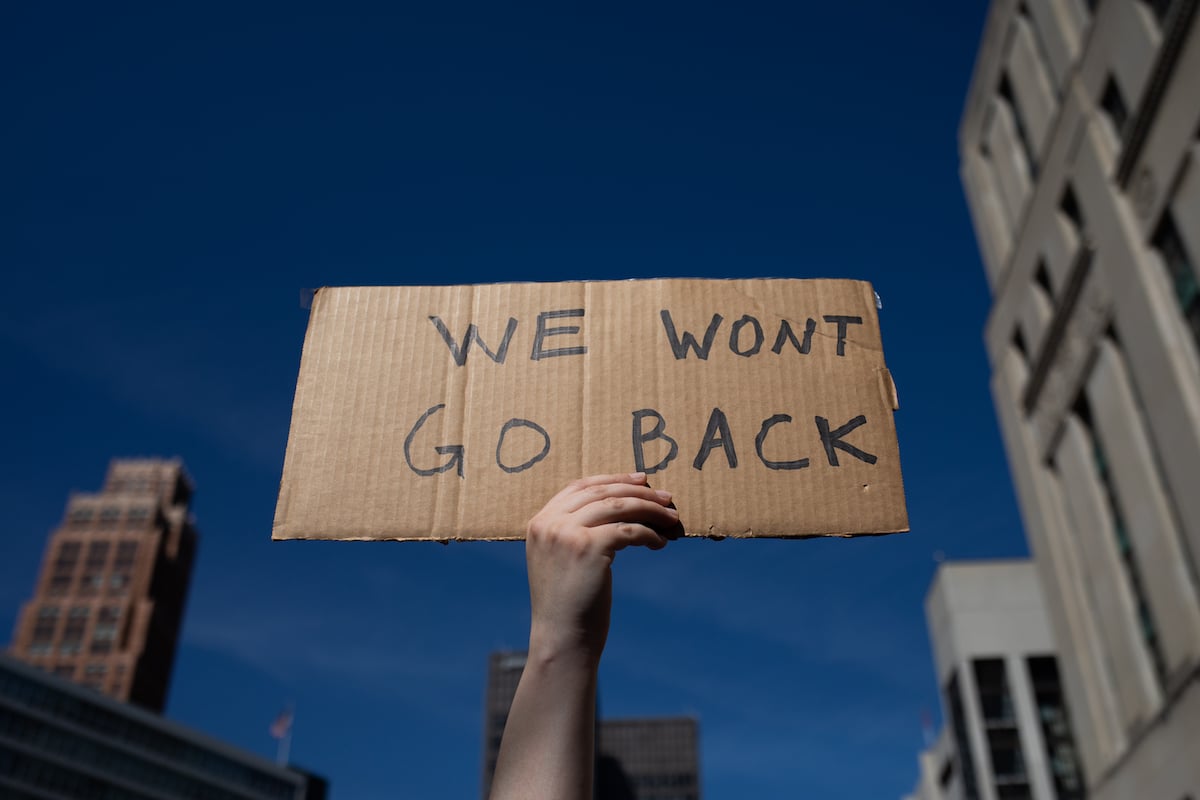 A hand holds up a small handwritten cardboard sign against a city sky background reading "We won't go back"
