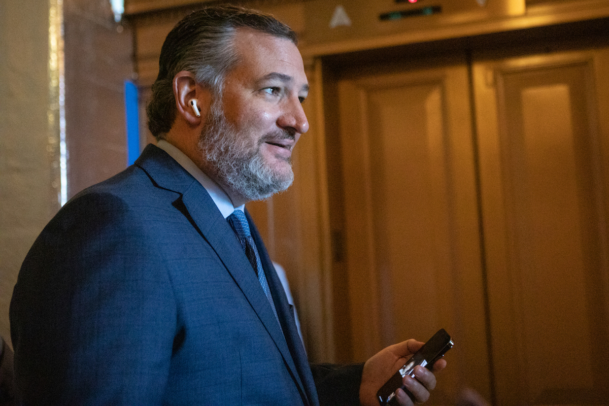 Ted Cuz smirks, holding his phone and wearing airpods in a Senate hallway