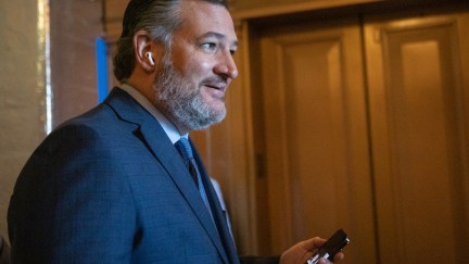 Ted Cuz smirks, holding his phone and wearing airpods in a Senate hallway