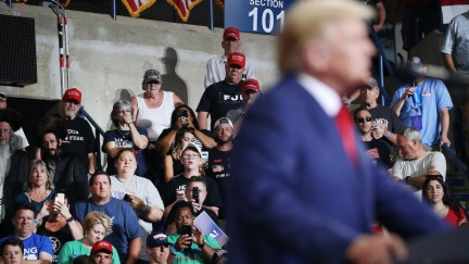 Donald Trump speaks from a podium at a rally with a crowd of supporters seen in focus behind him.