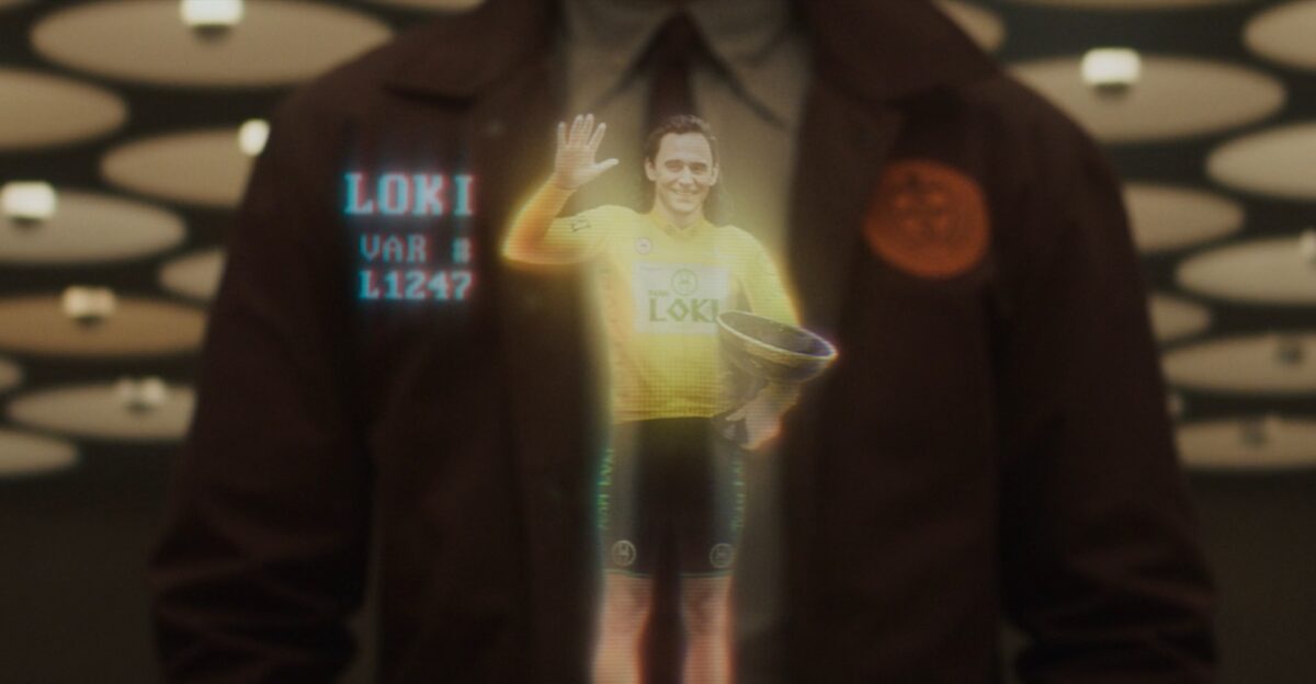 A hologram of Loki wearing a cycling uniform and holding a large trophy.