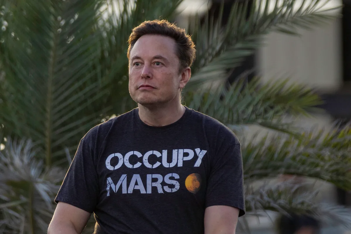 Elon Musk sits outside, staring blankly into the distance, wearing a shirt reading "occupy mars"
