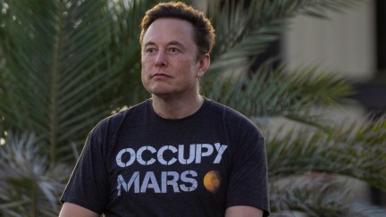 Elon Musk sits outside, staring blankly into the distance, wearing a shirt reading 