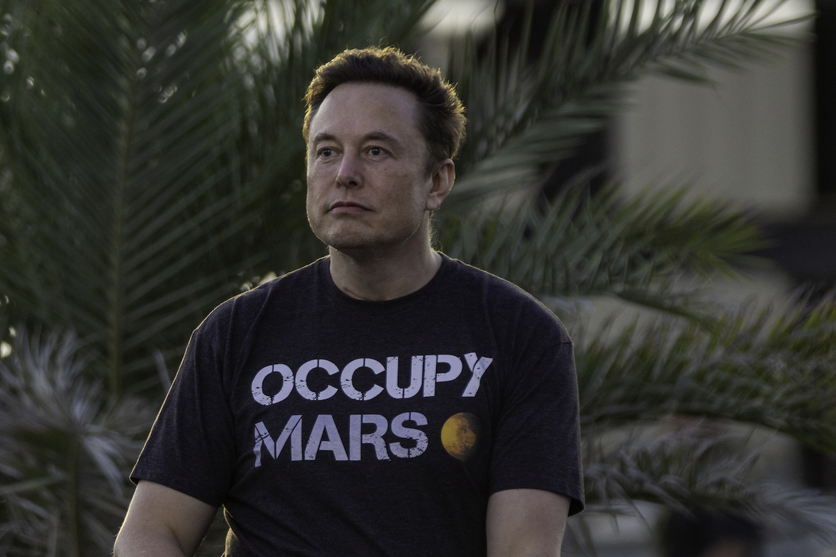 Elon Musk sits outside, staring blankly into the distance, wearing a shirt reading "occupy mars"