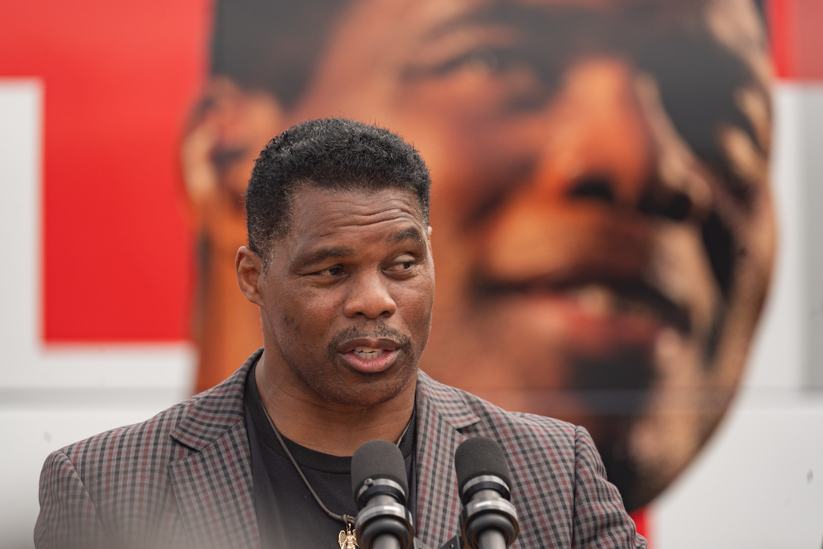 Herschel Walker speaks into microphones, standing in front of a giant picture of his face in the background
