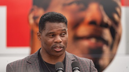 Herschel Walker speaks into microphones, standing in front of a giant picture of his face in the background
