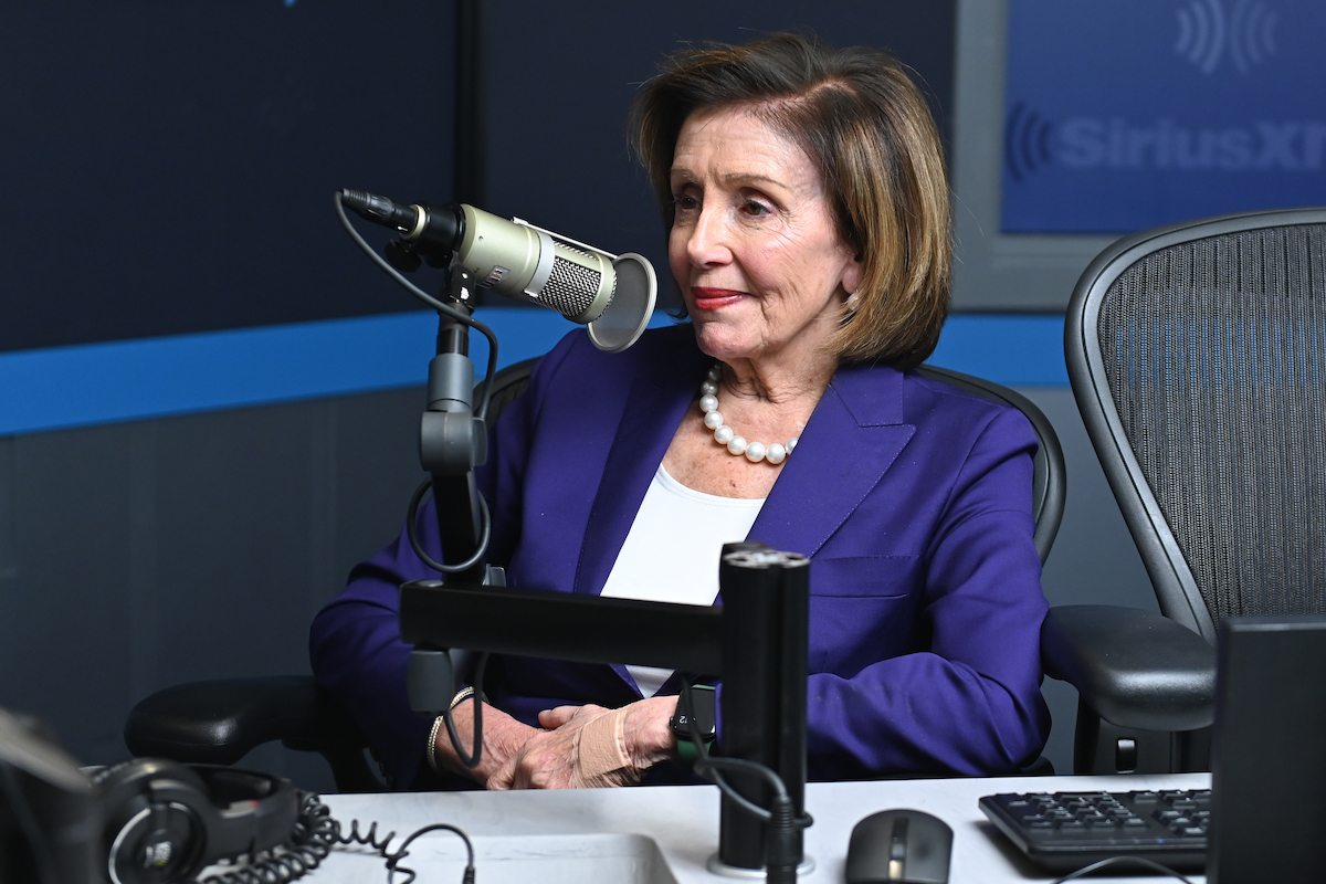 Nancy Pelosi leans back in her chair and smirks during a radio interview with a large microphone in front of her.
