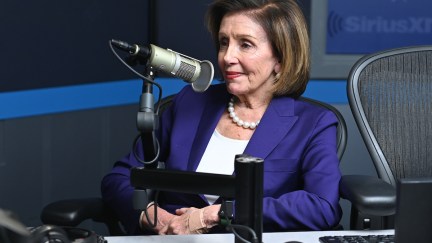 Nancy Pelosi leans back in her chair and smirks during a radio interview with a large microphone in front of her.