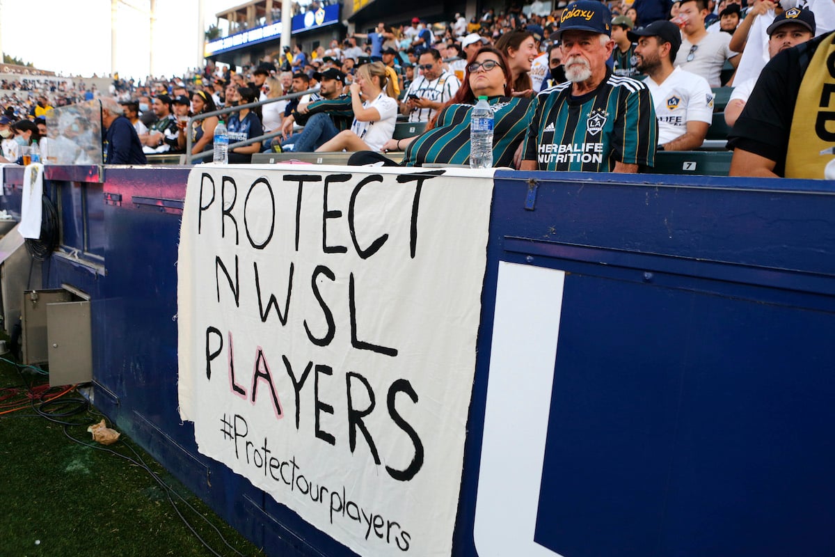A handwritten banner reading "Protect NWSL Players" hangs in the stands at a soccer game.