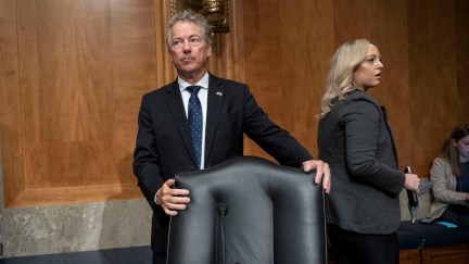 Rand Paul stands behind a chair, looking away nervously.