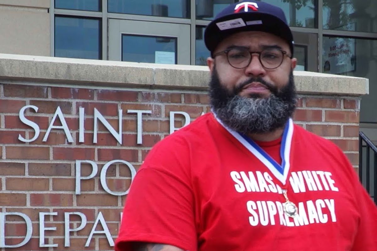 A Black man wearing a shirt reading "SMASH WHITE SUPREMACY" and a medal around his neck looks into the camera.