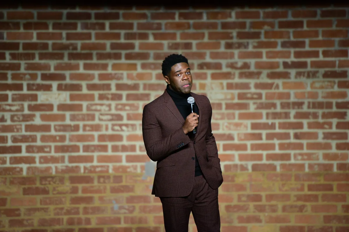 Sam Richardson stands in front of a brick wall holding a microphone.