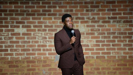 Sam Richardson stands in front of a brick wall holding a microphone.
