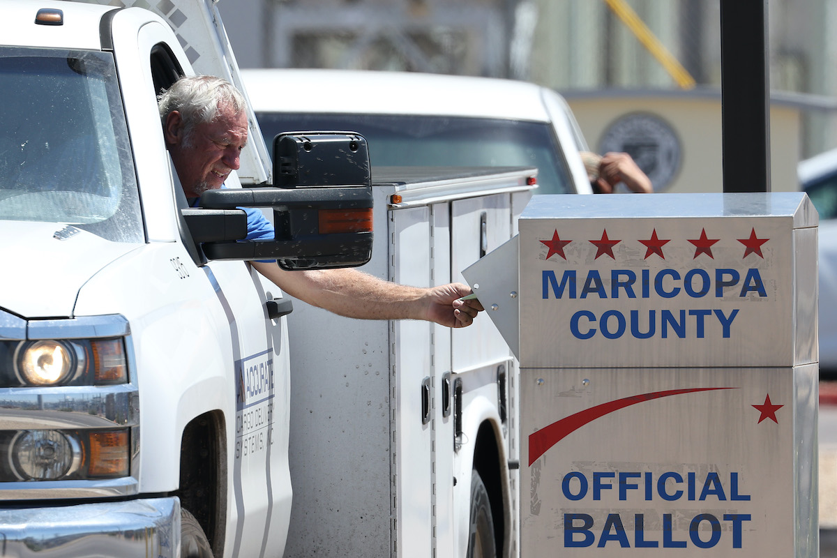 An older white man leans out of his truck window to drop a ballot in a Maricopa County official voter drop box.