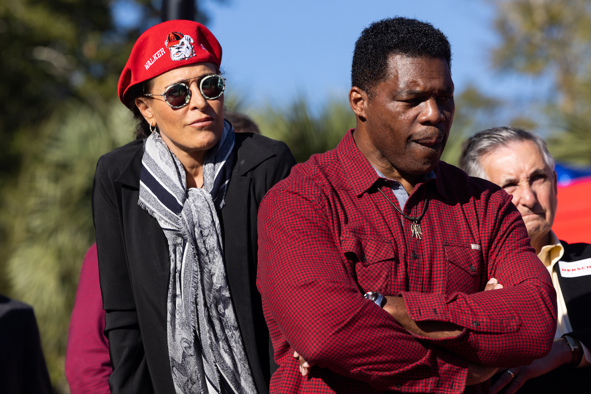 Herschel Walker stands with his arms folded, looking down while his wife stands behind him wearing a red beret with "Walker" and the image of a bulldog on it