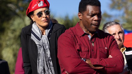 Herschel Walker stands with his arms folded, looking down while his wife stands behind him wearing a red beret with 