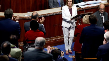 Nancy Pelosi wears a white suit and speaks from the House floor while members of Congress stand and clap.