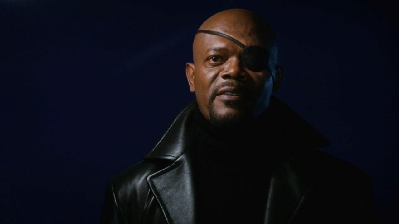 Nick Fury, in his iconic eyepatch and black coat, smiles against a black background.