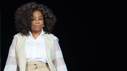 Oprah stands in front of a black background at a live event and smirks.