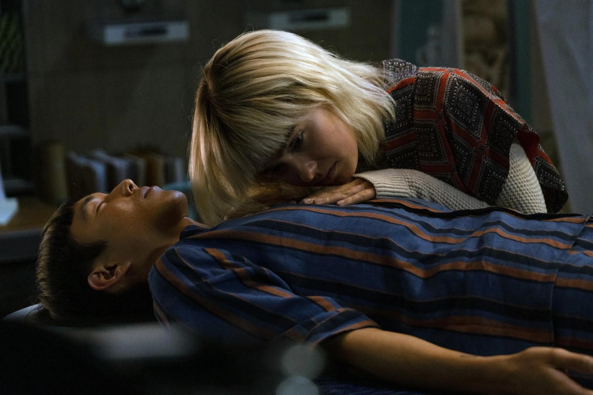 A girl standing over a boy who is reclining with his eyes closed, pressing her head against his chest, from "After Yang"