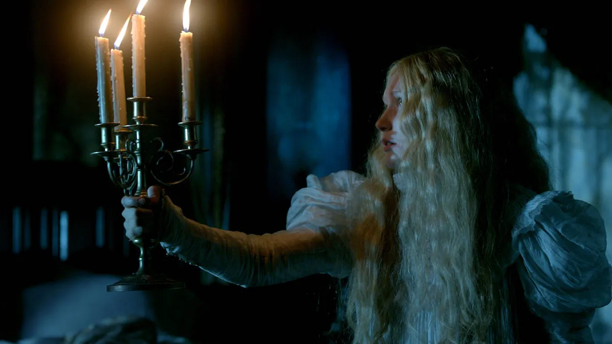 A woman (Mia Wasikowska) in white Victorian dress holds a candelabra to light her way down a dark hallway in a scene from 'Crimson Peak'