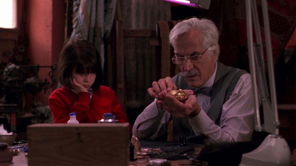An old man and a little girl are sitting at a table and examining a golden object in a scene from 'Cronos'