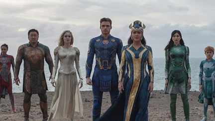 The cast of 'Eternals' stands on a beach in their superhero outfits.