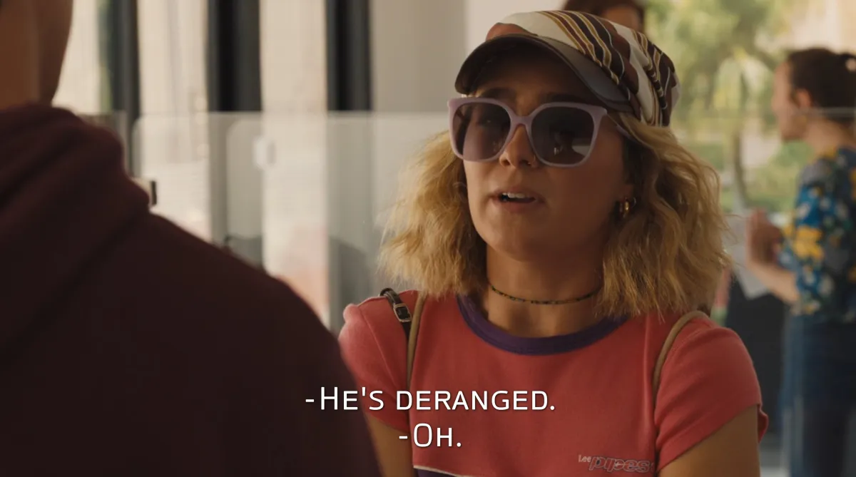 Portia wearing sunglasses and talking to Albie at the airport in 'The White Lotus.' The closed caption reads "He's deranged."