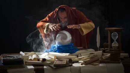 Fortune teller magician in fantastical smoky atmosphere using crystal ball. He is wearing a brown frock with hood. The background is black. Smoke is coming out from the desk. There are large amount of scrolls, few antique black books and a hourglass on desk.
