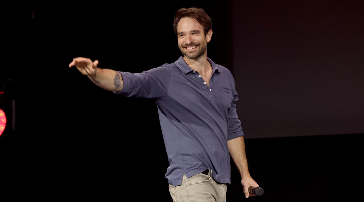 Charlie Cox waves to an unseen audience against a black background.