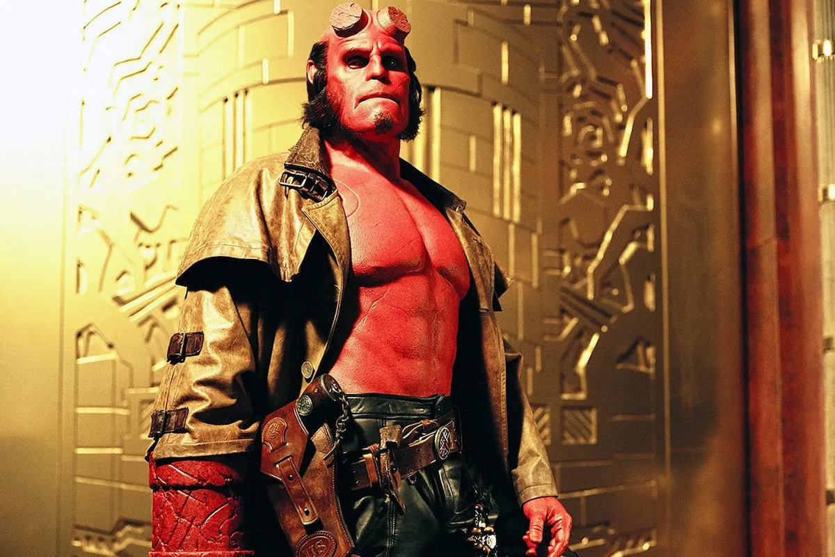 The comic book character Hellboy, portrayed by Ron Perlman, stands against a gold background and wears a trenchcoat. 