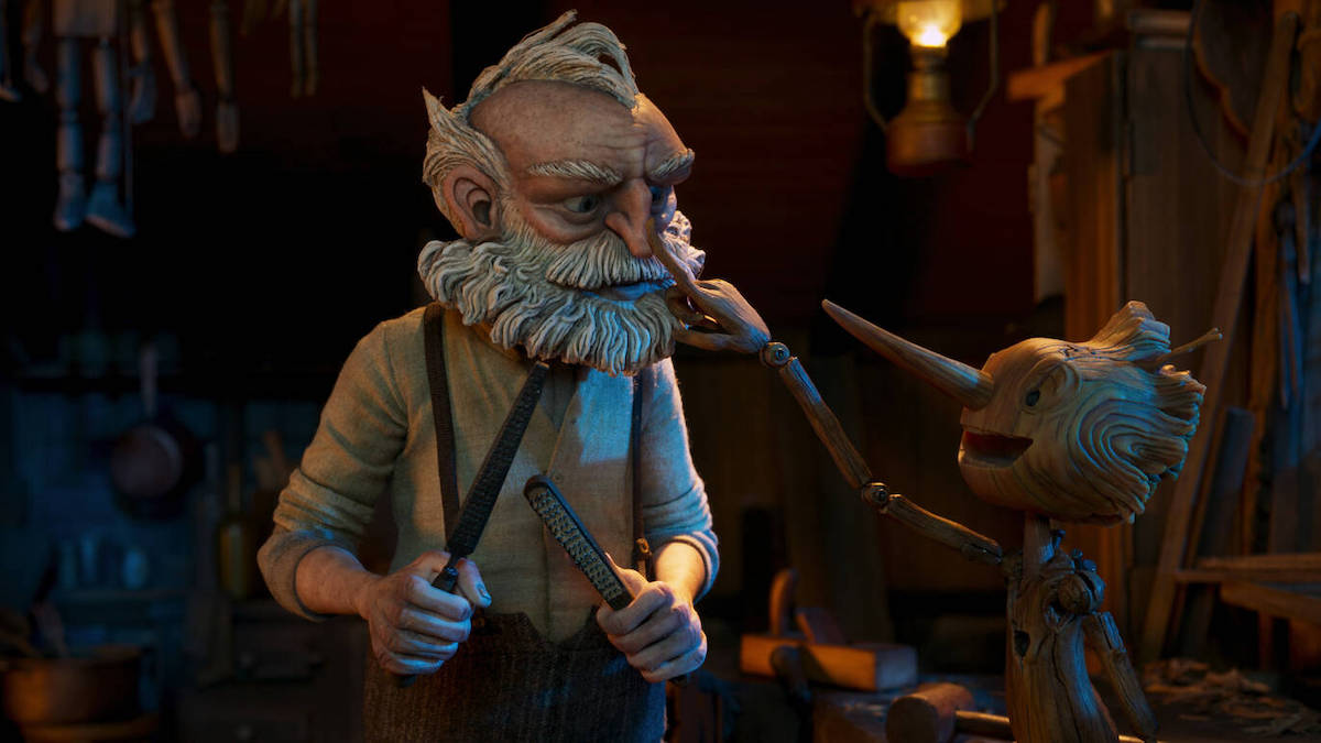 An old man holds woodworking tools while standing next to a wooden puppet seated on a workbench in Guillermo del Toro's 'Pinocchio'