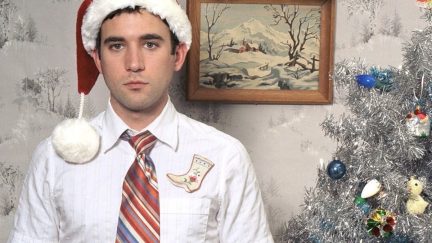 Sufjan Stevens wears a Santa hat and stands near a decorated Christmas tree