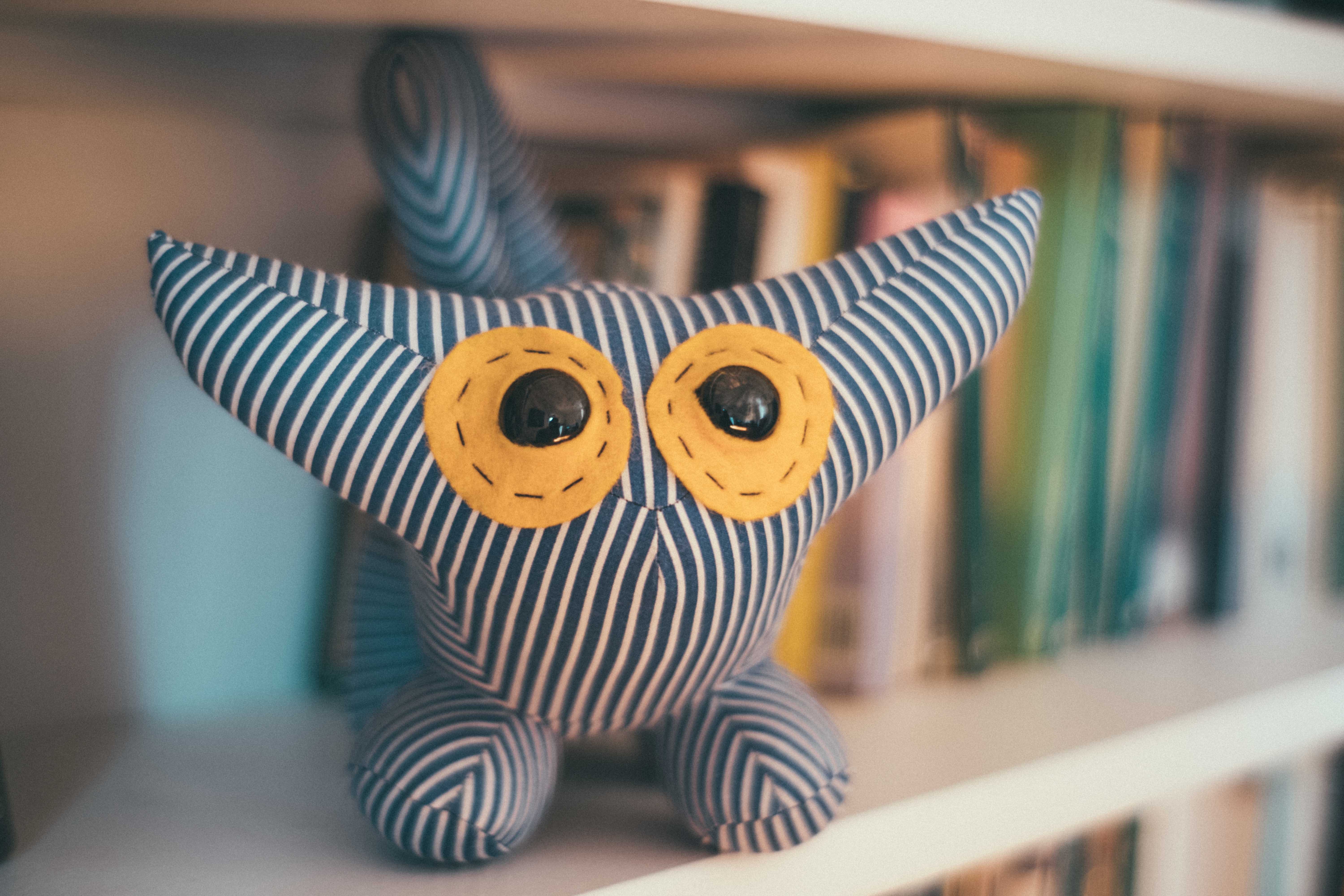 A stuffed animal with stripes, big ears, and big yellow eyes sits on a bookshelf. It's not clear what kind of animal the toy is meant to look like.