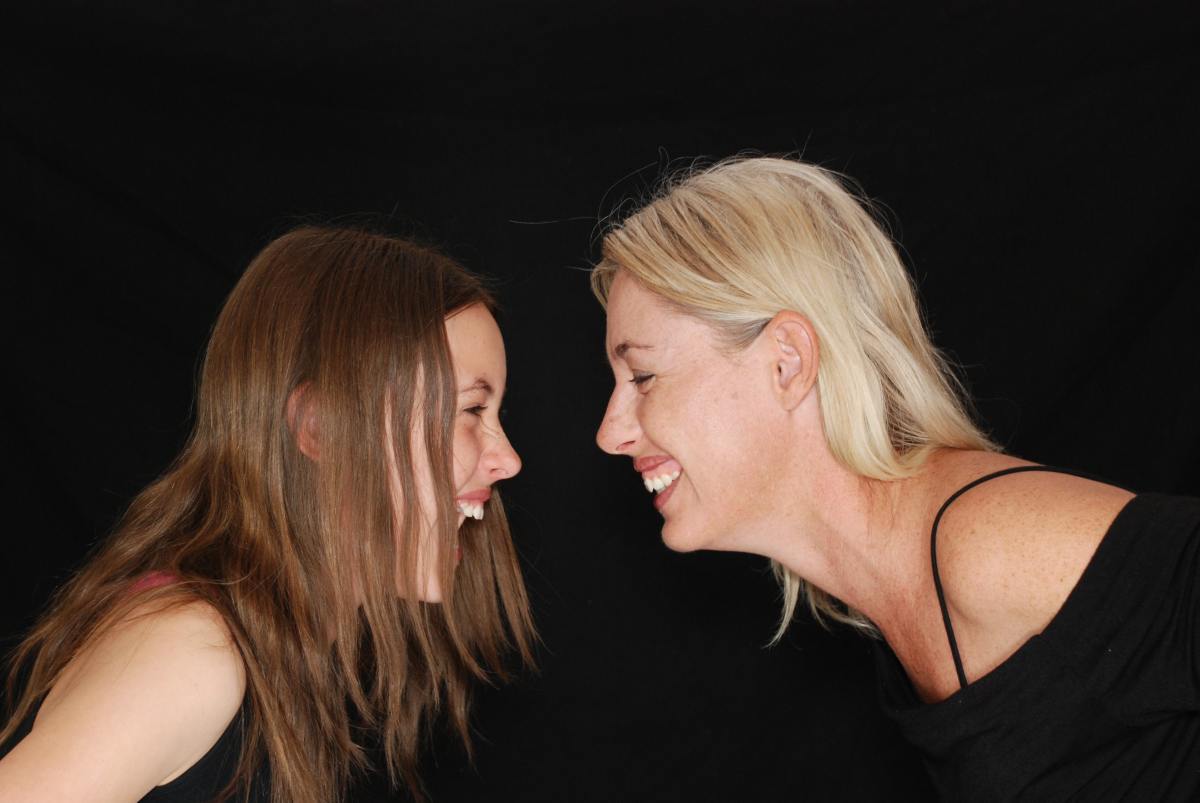 An older and younger woman smile at each other against a black background.