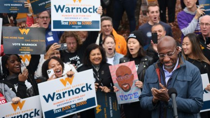 Raphael Warnock smiles and claps while standing in front of a group of supporters holding campaign signs.