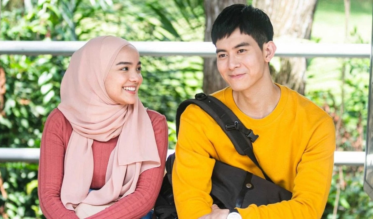 A smiling Malaysian woman in a tudung sitting on a park bench next to a smiling Chinese man wearing a cross body bag in a scene from 'Kongsi Raya'