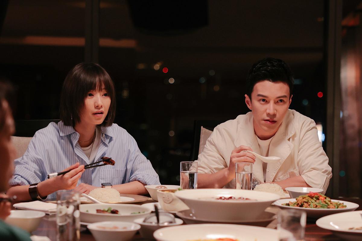 A woman on the the left and a man on the right, sitting at a table full of platters, eating in a scene from 'Reunion Dinner'