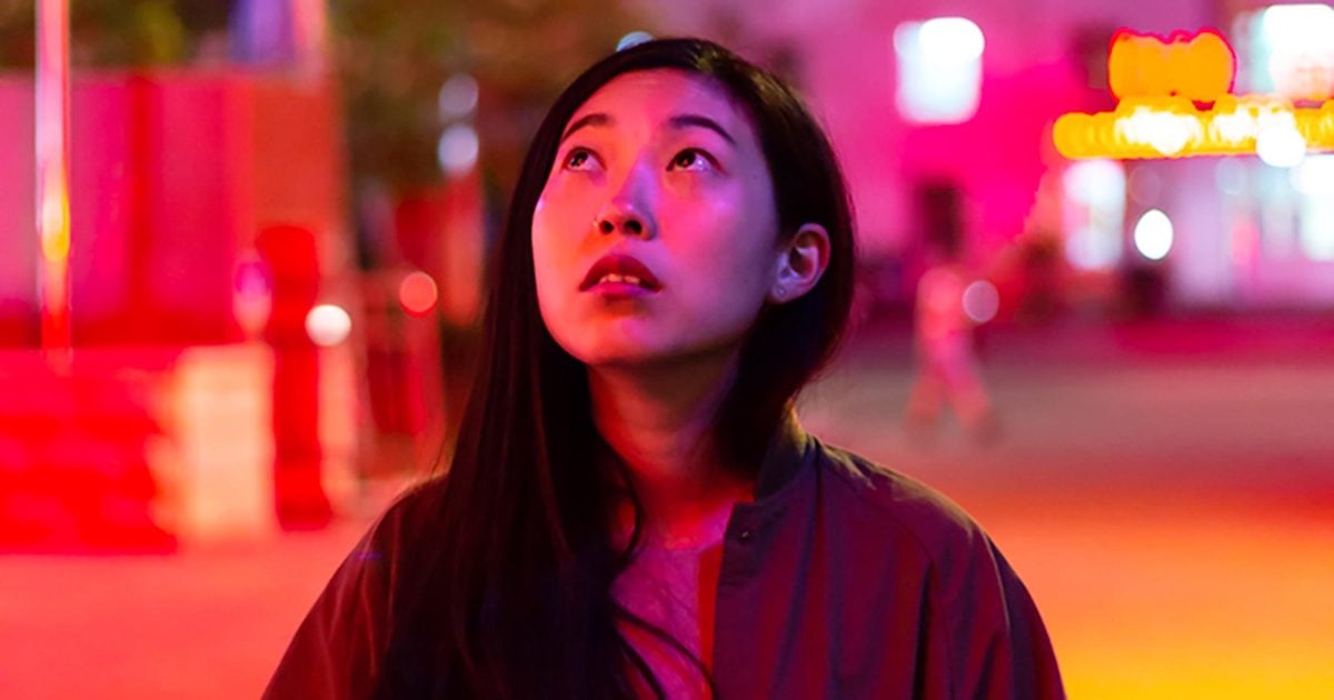 A close up of the Asian-American actor Awkwafina in the film The Farewell. She is lit by neon lights and looking up at the sky