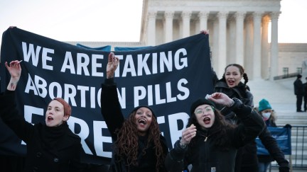 A group of women dressed in black in front of the US Supreme Court stand in front of a banner reading 