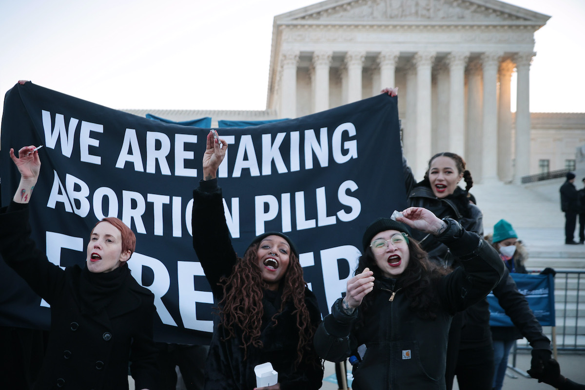 A group of women dressed in black in front of the US Supreme Court stand in front of a banner reading "we are taking abortion pills"
