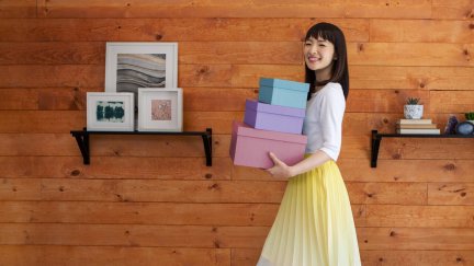 Marie Kondo smiles and holds three pastel-colored boxes against a wooden wall.