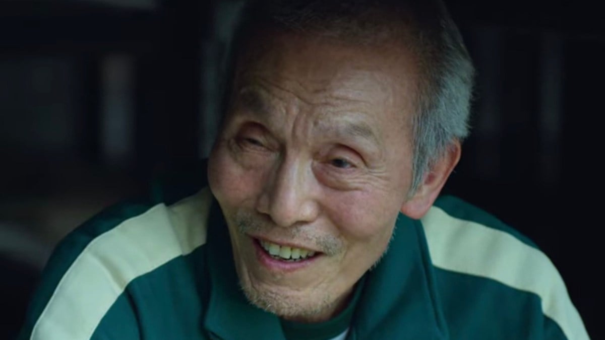 An elderly Asian man in a green and white tracksuit jacket softly smiling.