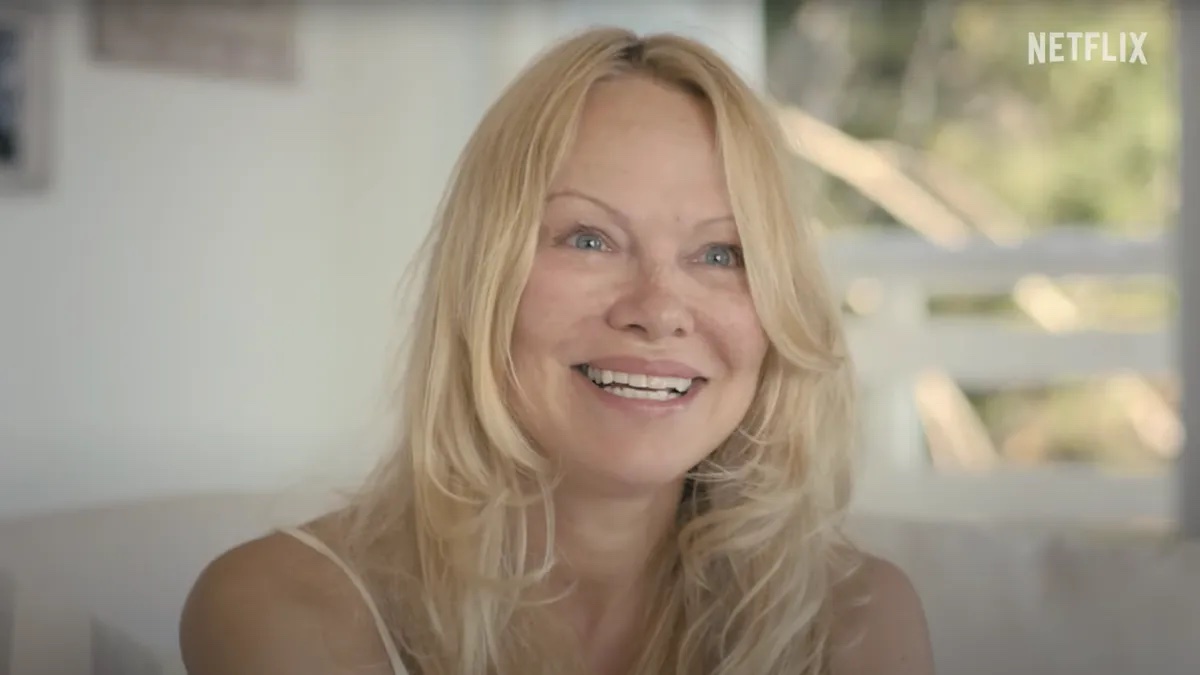 Pamela Anderson smiles without makeup on, sitting on a porch.